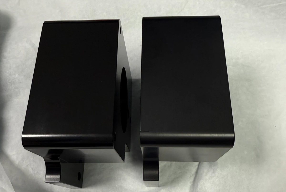 Two black anodized aluminum CNC parts showing visible shade and gloss variation under similar lighting